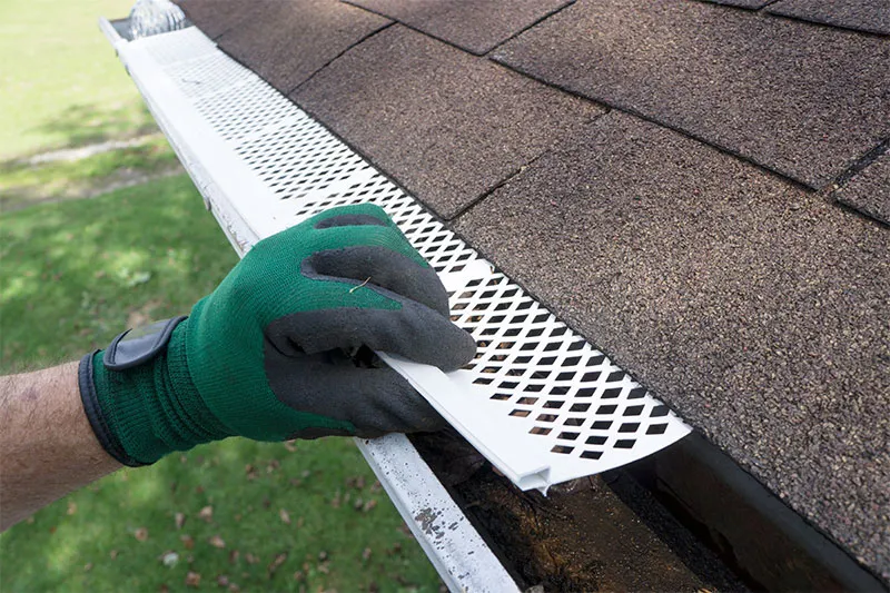 Gloved hand fitting a perforated gutter guard over a gutter at the edge of a shingle roof
