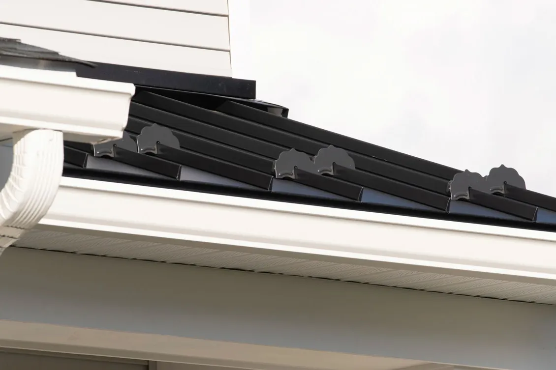 White gutter and downspout under a dark roof edge beside a stone wall