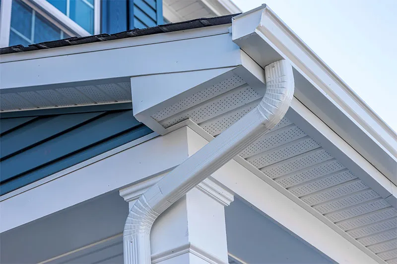 White K-style gutter and downspout below vented soffit on a house with blue siding